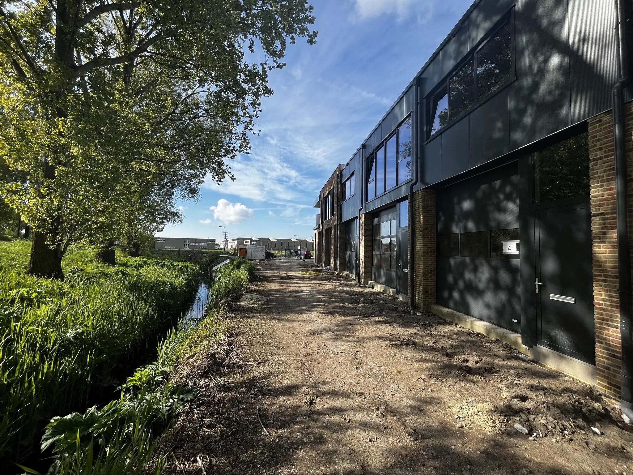 Bedrijfsgebouwen aan de Broekdijk West 19-4 met een onverharde weg en een sloot langs een groene berm.